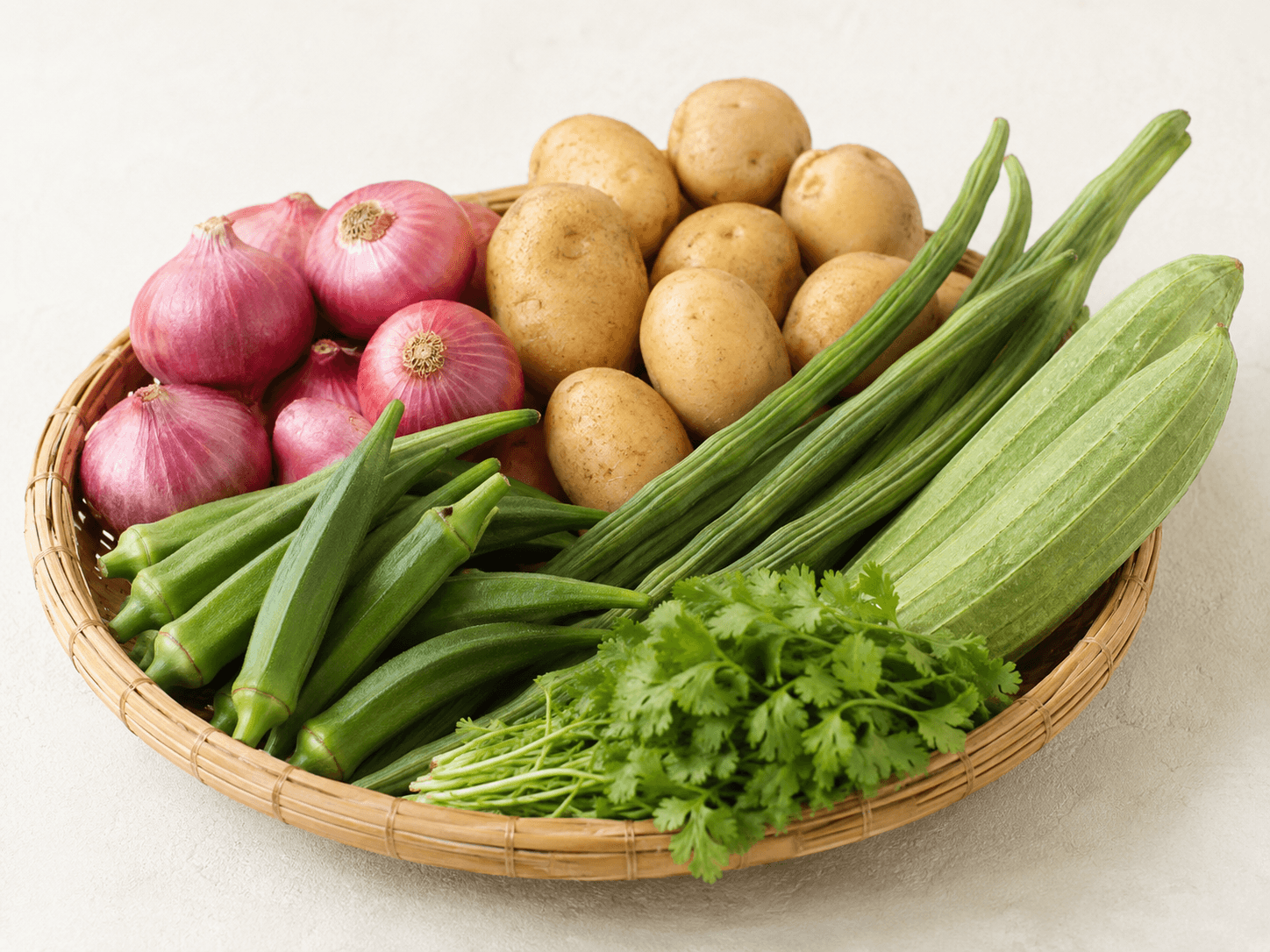 Editorial still life of premium hand-graded Indian fresh vegetables — a bundle of bright green okra, a handful of fresh drumstick (moringa) pods, smooth ridge gourd, and fresh coriander — arranged in a shallow woven basket on a clean off-white surface, soft natural daylight.