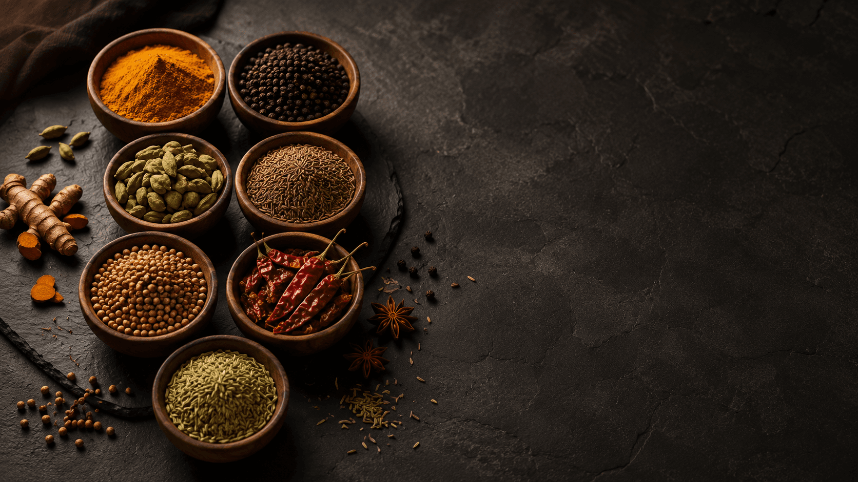 Editorial still life of premium Indian spices — turmeric, black pepper, cardamom, cumin, coriander, red chilli, fennel — in small wooden bowls on a dark slate surface.